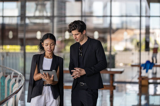 Young business officers in black jacket exchange ideas on business using tablet to provide data and information at a terrace outside of the building. Conversation of two coworkers in casual dress
