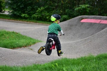 Toddler boy wearing helmet rides red balance bike on asphalt pump track. Active child learning...