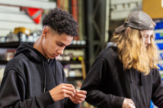 Young mechanic holding small mechanical part beside colleague near workbench