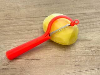 Peeled potato with red peeler on wooden table, close-up of kitchen utensil and vegetable prepared...