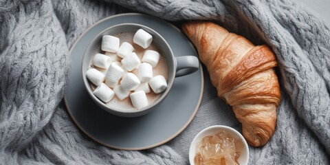 Cozy morning treat a mug of hot chocolate with marshmallows, a croissant, and a small bowl of jam, all resting on a grey knitted blanket