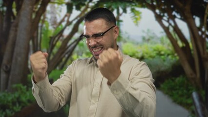 Young hispanic man wearing glasses raises clenched fists and grins broadly in green park; triumph celebration joy.