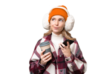 Young English woman wearing winter muffs over isolated background holding coffee to take away and a mobile while thinking something