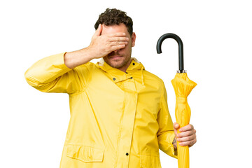 Brazilian man with rainproof coat and umbrella over isolated chroma key background covering eyes by...