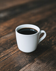 A simple white cup filled with black coffee sits on a rustic wooden surface, exuding warmth and comfort in a cozy setting.