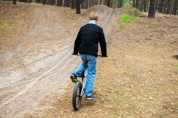 Rear view of a young person in a black jacket and jeans, riding a BMX bicycle uphill on a dirt path...