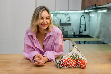 Smiling woman in pink shirt leaning on kitchen counter next to mesh bag with fresh vegetables. Healthy lifestyle and eco-friendly food shopping at home.