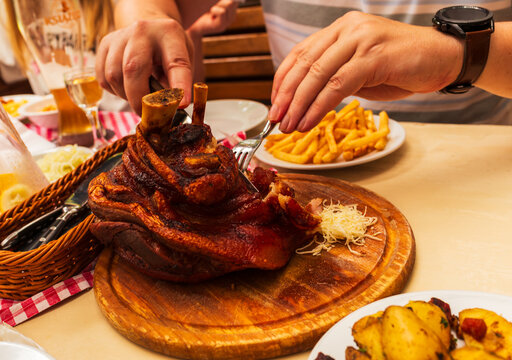 Close view of hands slicing a succulent roasted pork knuckle (golonka) served with sauerkraut, potatoes, and beer in a rustic restaurant. The caramelized crust and steam convey hearty Central European