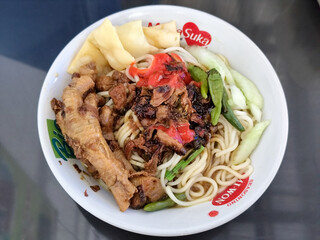 A complete bowl of chicken noodles with claws, dumplings, chicken pieces and red chili sauce on top, traditional Indonesian food.