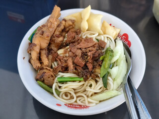 A complete bowl of chicken noodles with claws, dumplings, chicken pieces and red chili sauce on top, traditional Indonesian food.
