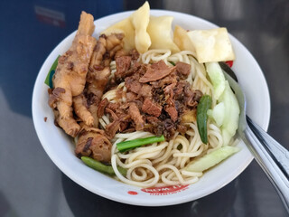 A complete bowl of chicken noodles with claws, dumplings, chicken pieces and red chili sauce on top, traditional Indonesian food.