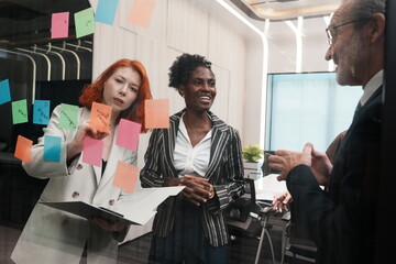 group of employees and an elderly male executive and two multinational female team members stand together, brainstorming and debating a strategic plan using stickers on a glass wall in conference room