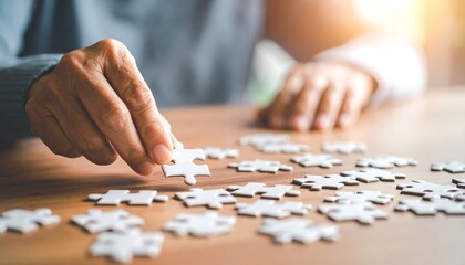 A focused person's hand meticulously places a puzzle piece onto a wooden table, filled with scattered puzzle pieces.