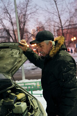 A man in a winter coat inspects the engine of a car at dusk. Snow blankets