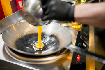 Chef pours beaten eggs into hot wok on electric stove, wearing black glove. Professional kitchen...