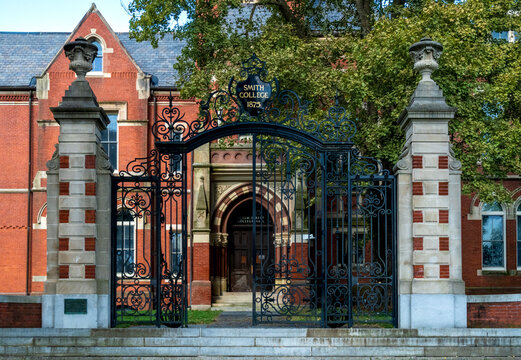 Northampton, MA - US - Aug 24, 2025 Ornate black iron gates and stately stone pillars frame the entrance to a historic red-brick College Hall at Smith College, nestled among lush trees.