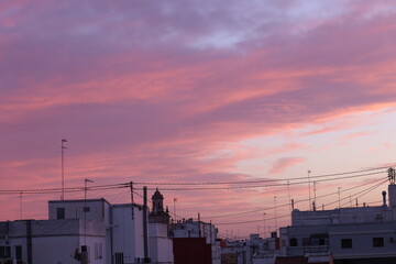 A vibrant Spanish sunset with pink and purple clouds over white rooftops and a church dome, framed by antennas and power lines.