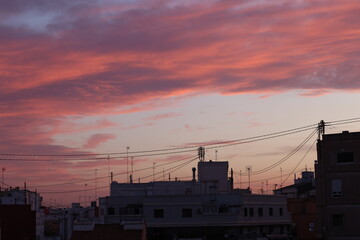 A vibrant Spanish sunset with pink and purple clouds over white rooftops and a church dome, framed by antennas and power lines.