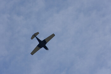 A small single-engine airplane flies high against a cloudy blue sky, captured in a dynamic upward view. The image evokes a sense of freedom, travel, and aviation, with a serene and expansive mood.