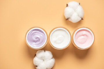 Set of cosmetic creams of different colors in open jars with fluffy cotton flowers on yellow pastel background, top view, anti-aging cream, moisturizing cream