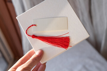 Female hand holding a white luxury card mockup with a red tassel against a softly lit background. The card is ready for a message or decoration, minimalist design.