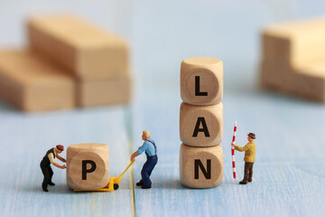 Miniature worker figure pulling a wooden cube with the letter P to stack with L, A, and N blocks, forming the word PLAN. Business concept for strategy, planning, teamwork, and goal achievement.
