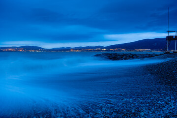 Storm in Gelendzhik on long exposure. Waves in form of fog run into beach of Gelendzhik. Twilight, sunset. Mountains and city lights on horizon