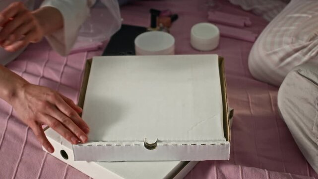 Zoom out shot of group of young cheerful women wearing pajamas and eye patches enjoying bachelorette party at home, sharing pizza and drinking champagne while sitting on pink bed - Powered by Adobe