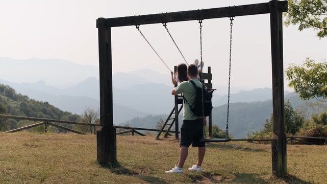 Real-time medium-wide shot of a happy father gently pushing his smiling daughter on a wooden swing with a mountain view.