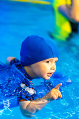 Child learning to swim in a pool. A young child in a blue swimming cap practices swimming in a pool while showing excitement and determination.