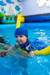 Child learning to swim in a playful pool. A young child in a blue swim cap practices floating with...