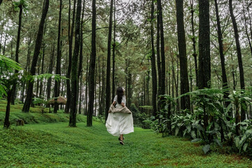 A woman in a white dress and brown sweater runs through a pine forest, hair flowing freely,...
