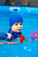 Child playing in pool. A young child in a blue cap swims joyfully in a clear pool, playing with colorful toys on a sunny day.