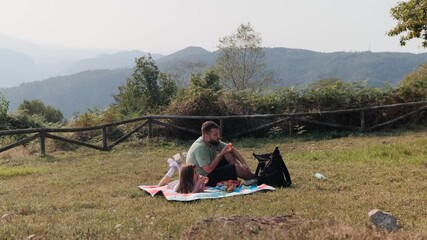 Real-time candid shot of a father and young daughter lying on a picnic blanket and sharing snacks on a sunny mountain meadow.