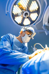 Surgeon performing surgery. A surgeon, dressed in blue scrubs and a mask, focuses intently on a...
