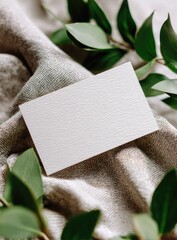 Blank textured card rests on neutral fabric, surrounded by lush green foliage; minimalist, elegant aesthetic