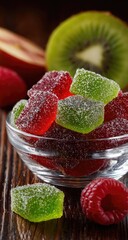 A glass bowl filled with vibrant red and green sugared jelly cubes sits on a wooden surface, alongside fresh raspberries, a kiwi, and apple slices