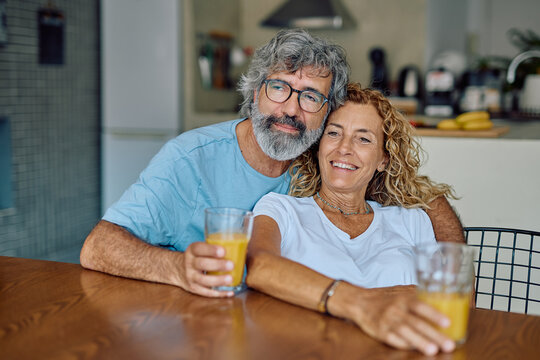 Senior couple enjoying refreshing orange juice together in a modern kitchen, embracing and sharing joyful smiles during a cozy breakfast - Powered by Adobe