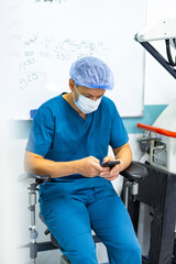 Doc checks phone on break. Healthcare worker in scrubs and mask sits on chair looking at phone, surrounded by medical equipment and notes.