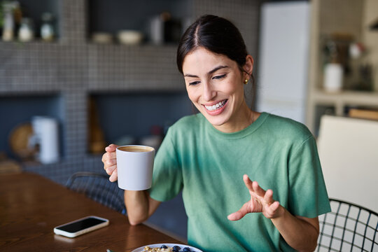 Portrait of a young woman using tablet or laptop computer, having online meeting or browsing internet, drinking coffee eating breakfast, drinking juice  in the kitchen at home