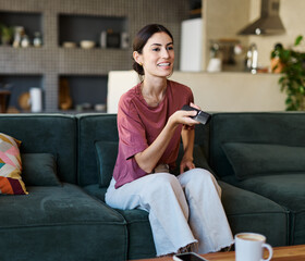 Obraz premium Portrait of a young latina woman having fun holding a remote control and watching tv at home