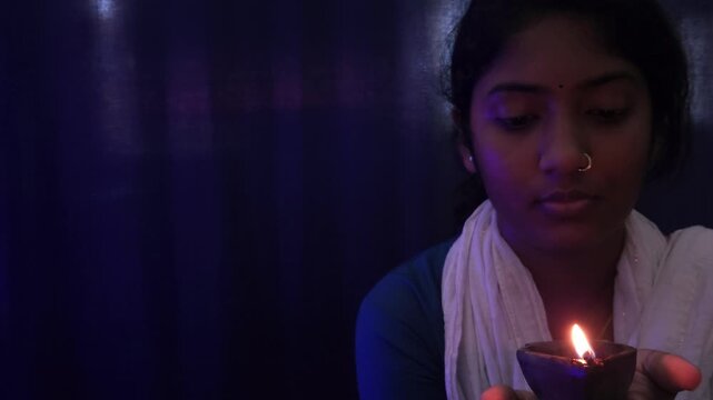 Indian teenage girl&rsquo;s hands gently hold a glowing diya lamp, radiating warm light and symbolizing celebration. The soft bokeh lights evoke a serene and festive Diwali atmosphere.