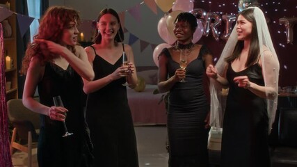 Group of young women in elegant dresses and bride-to-be in veil holding champagne glasses and dancing during festive bachelorette party in decorated room