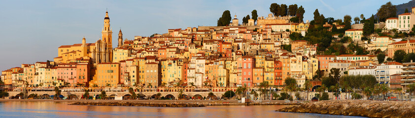 Panoramic sunset view of Menton, France, with colorful old town buildings glowing in warm light, set against the Mediterranean Sea and blue sky on the French Riviera