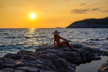 Brunette in pink bikini with straw hat at seaside during sunset in Montenegro