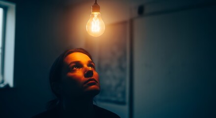 Pensive woman gazes at a glowing lightbulb in dimly lit room with dark walls, concept for inspiration, problem solving and eureka moment