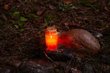 A glowing red memorial candle rests on a stone in the forest, casting warm light and symbolizing...