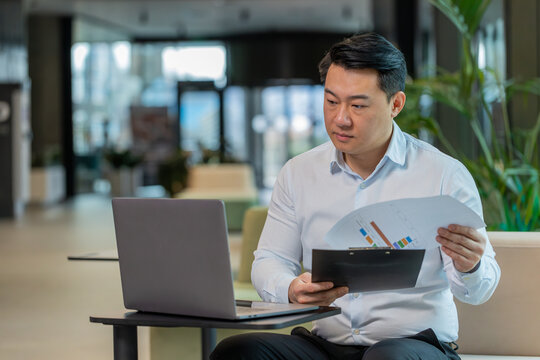 Chinese businessman working with documents, analyzing information, reading papers prepare financial report. Young male entrepreneur in formal using laptop analyzing graph documents in office lobby.