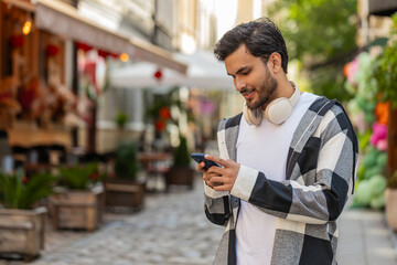 Indian young man using smartphone typing text messages in social media application online, surfing internet, relaxing, taking a break outdoors. Hispanic guy tourist standing on urban city street.