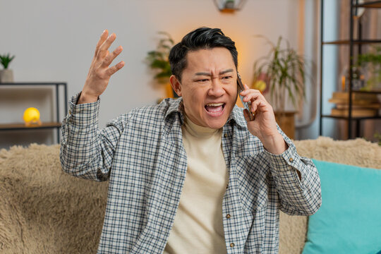 Angry young man arguing on smartphone while sitting on couch at home. Quarrel. Chinese mature guy talking loudly and shouting into cellphone, maliciously conveying and proving point through gestures. - Powered by Adobe